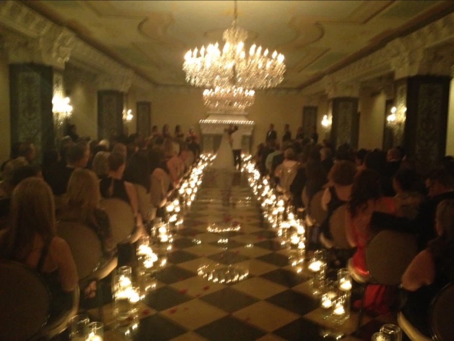 Bride walking a candlelit aisle in a chandelier-lit ballroom ceremony